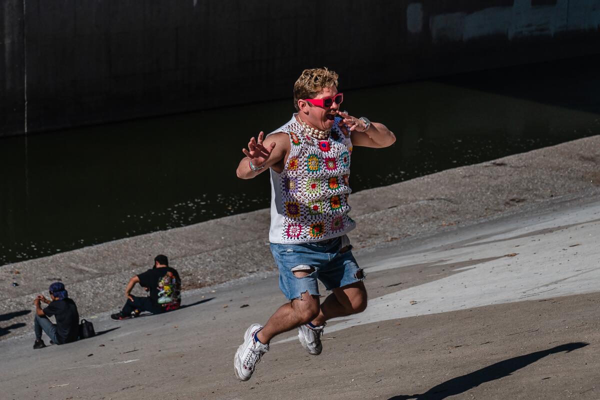 A man in red sunglasses and a flowery tank top jumps on the concrete embankment at the river.