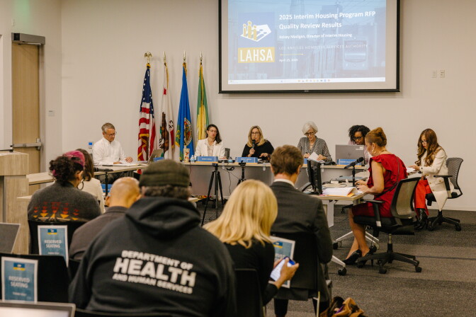 Panelists sit at a table in front of a screen during a public meeting, a man with a hoodie labeled Department of Health and Human Services is among those in the audience.