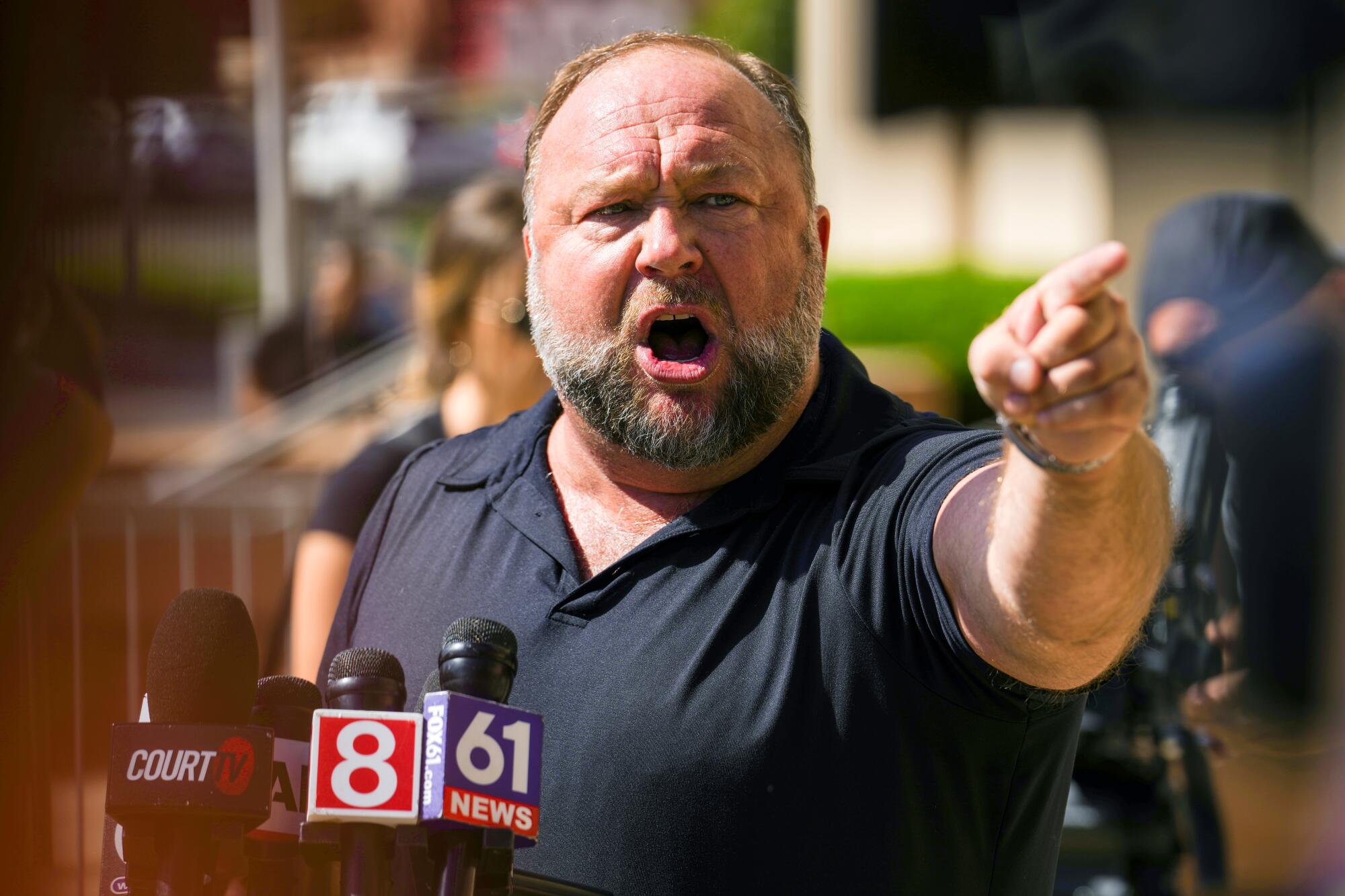 A balding man with a beard in a black collared shirt stands near a set of microphones and points.