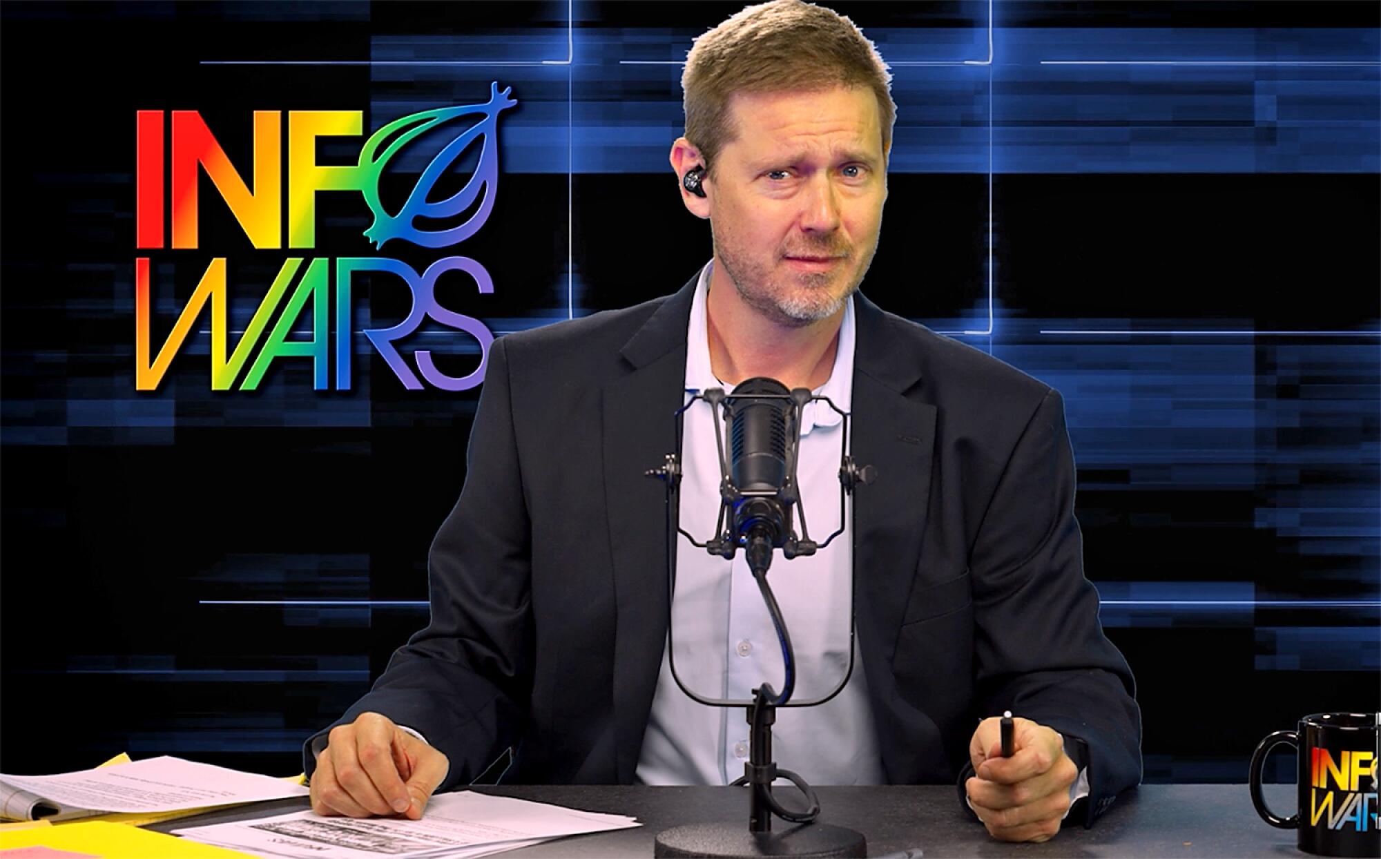 A man in a blazer and shirt sits at a desk with a microphone in front of him. The InfoWars logo is displayed on his left.