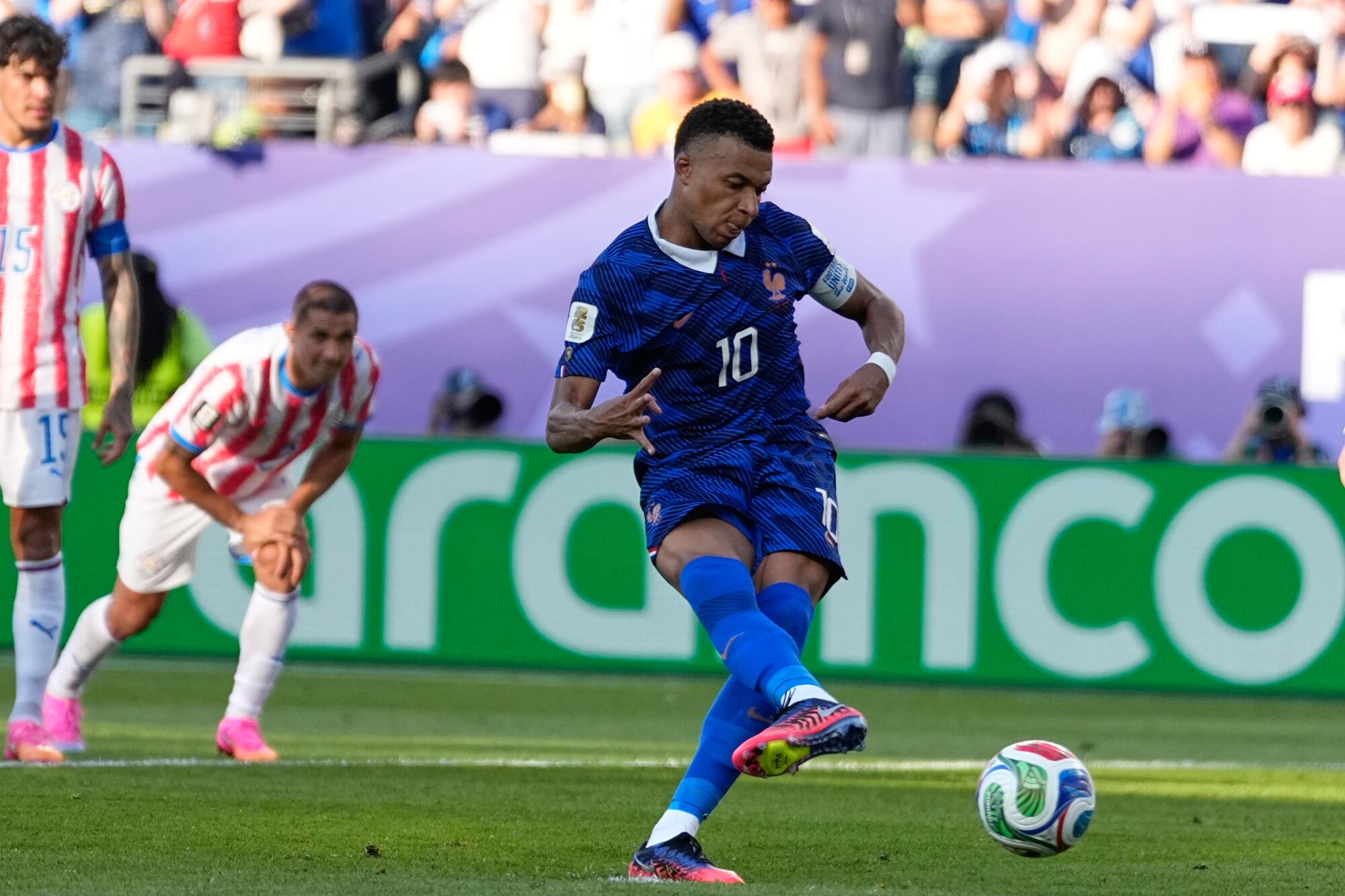 A soccer player in a blue France uniform kicks the ball as another player in a red and white Paraguay uniform watches