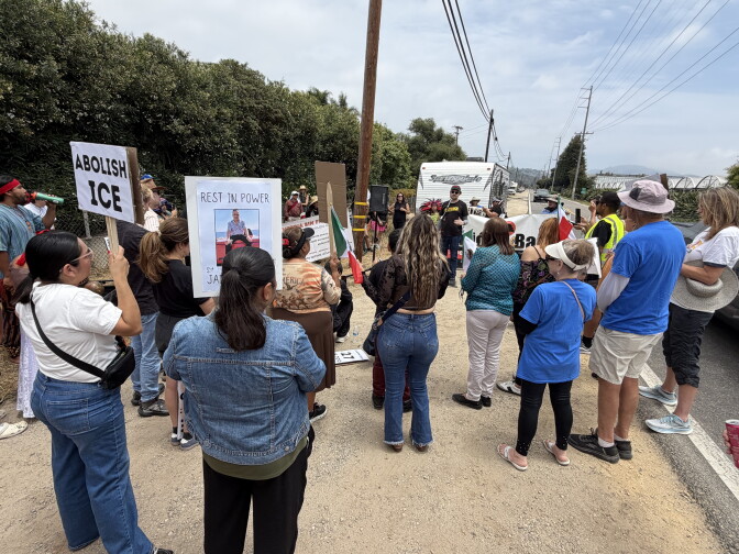 Crowds of people are standing facing away from the camera and looking towards a speaker. They are huddled in a circle with some holding fans, many are wearing hats and sunglasses.