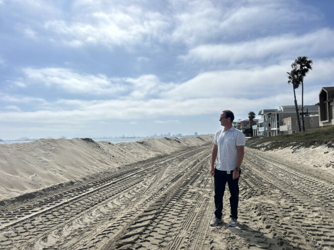 A middle aged man with light skin and a white collared T-shirt and dark pants stands on a sandy beach looking at tall sand berms. Behind him are houses.