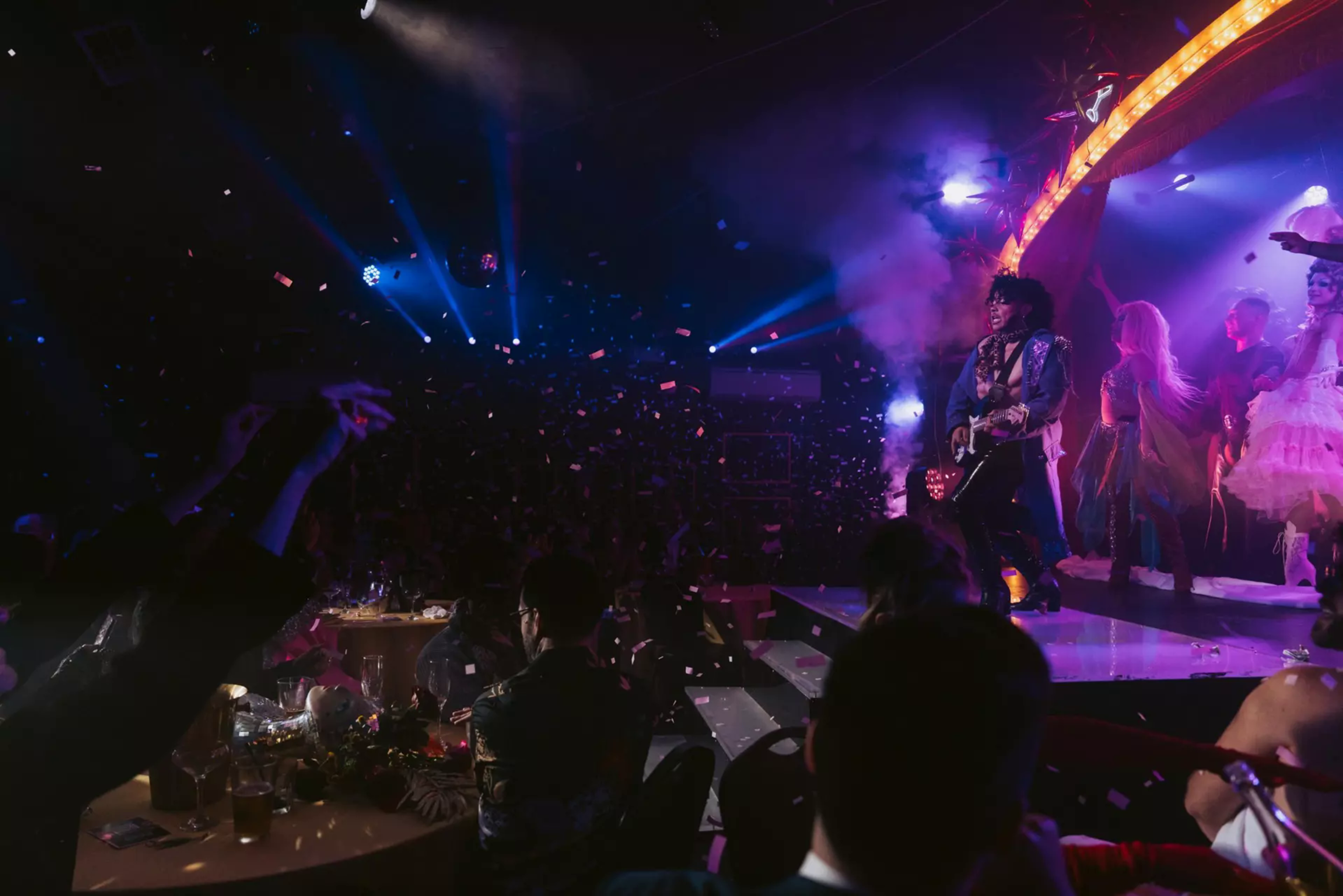 A lively concert with a guitarist on stage, colorful lights, confetti in the air, and an audience enjoying the performance at dimly lit tables.