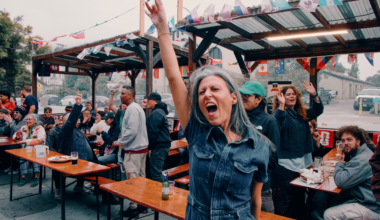 A lively crowd sits and stands around outdoor picnic tables under a decorated canopy, with a woman in front raising her hand and shouting joyfully.