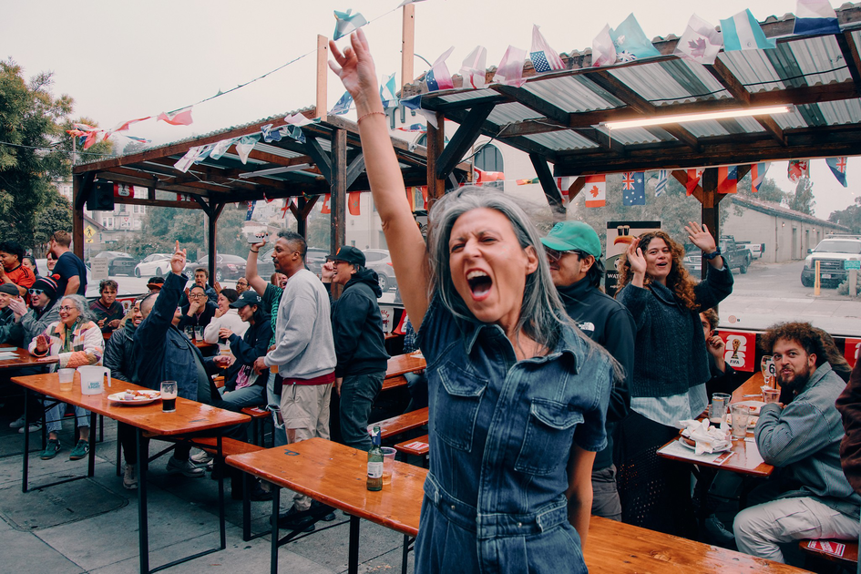 A lively crowd sits and stands around outdoor picnic tables under a decorated canopy, with a woman in front raising her hand and shouting joyfully.