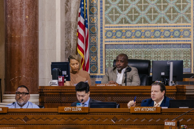 Various people sit from behind a wooden dais with wooden name tags that read "City Clerk" "City Attorney" and "Harris-Dawson." In the top chair is councilmember Marqueece Harris-Dawson, president of the council.