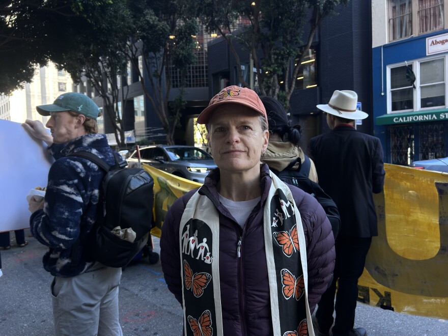 A woman with an orange baseball cap with a monarch on it, in a puff coat with a stole with monarchs on them stand on the sidewalk in San Francisco. Behind her are two people holding banners.