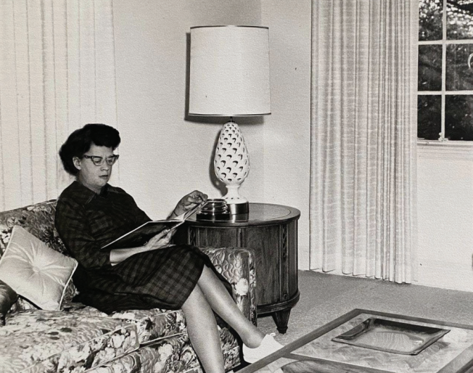 A female presenting woman sits on a couch and reads a magazine. She wears a checkered dress.