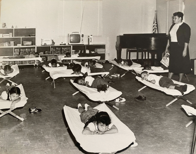 In a black and white photo, nine children sleep in foldable cots.