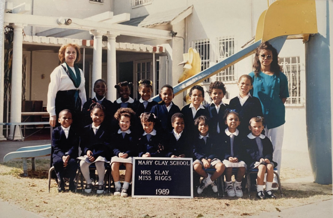 Fifteen children pose for a school photograph outdoors, in front of a building. Two female presenting adults stand on either side of the group.