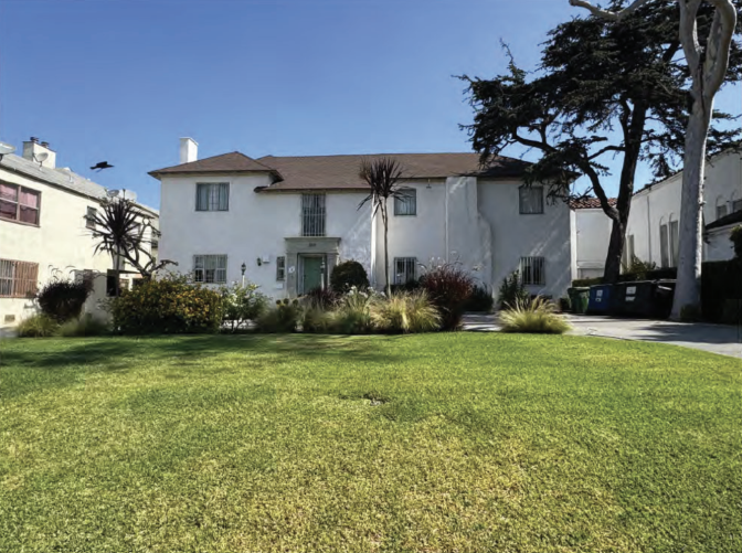 A two story, white building with a green lawn and trees around it, against a blue sky.