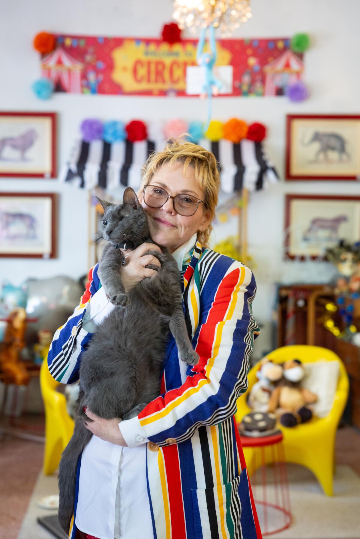 A woman with a blonde mullet hairsyle and a yellow, red, white, and blue blazer holds a one-eyed grey cat.