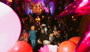 A crowded party with people socializing under chandeliers and colorful draped fabric, with balloons and decorations in the foreground.