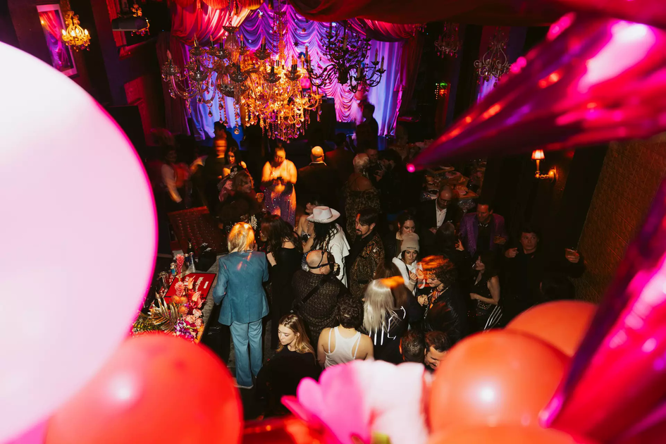 A crowded party with people socializing under chandeliers and colorful draped fabric, with balloons and decorations in the foreground.