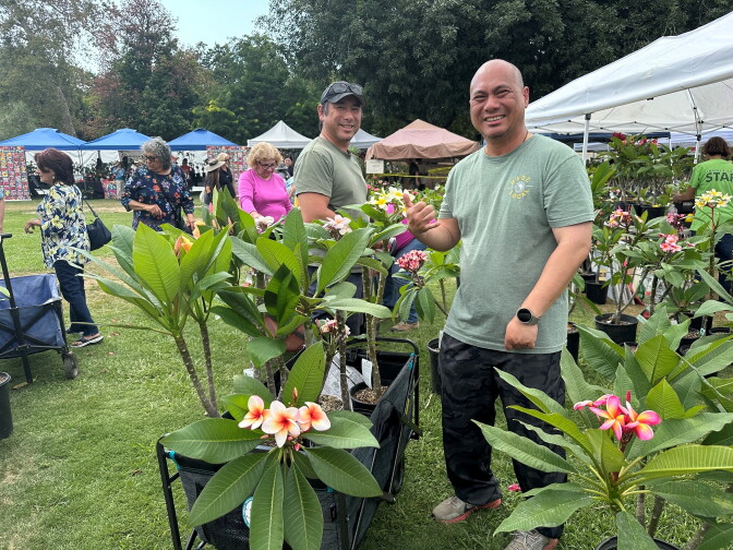 A medium-light-skinned man in a green t-shirt smiles in the foreground next to several plumeria plants, while other shoppers mill about behind him.