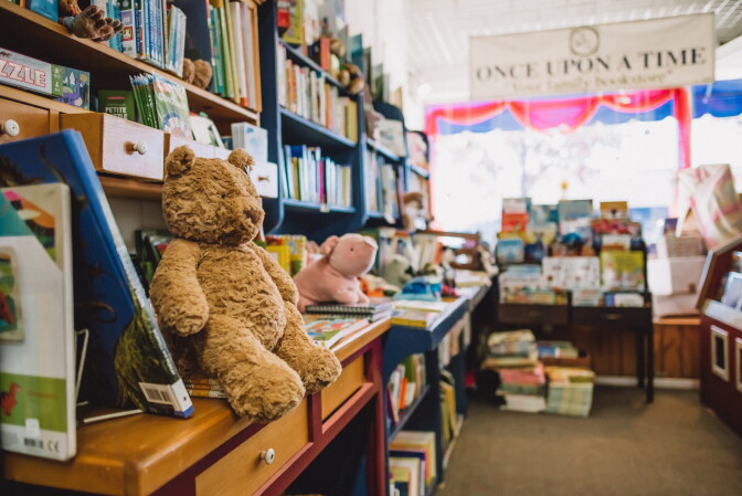 A children's bookstore is seen. There are stuffed animals, a pig and a teddy bear alongside a shelf with picture books and other children's books.
