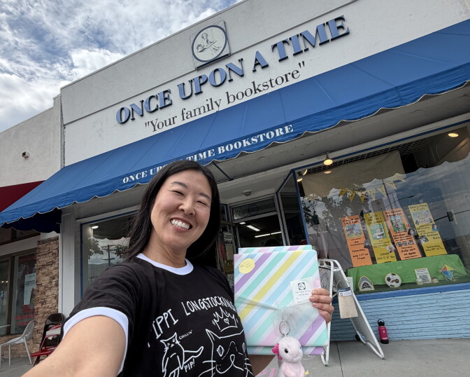 A woman stands in front of Once Upon A Time "Your family bookstore". She wears a black t-shirt and holds a gift wrapped present.