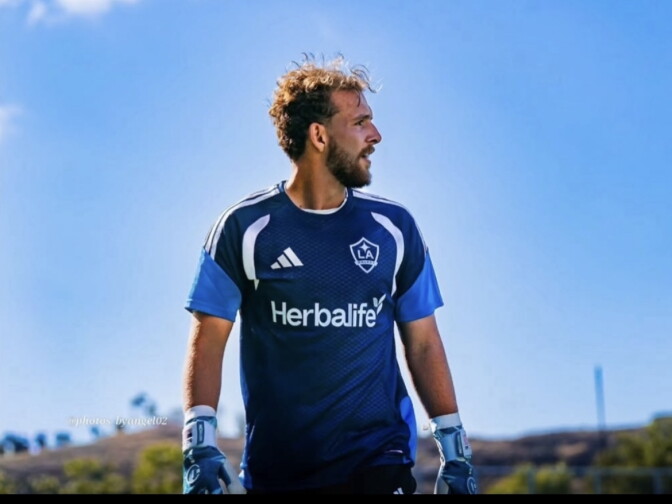 A light skinned male presenting person looks to the right. He wears a blue soccer team jersey and goal keeper gloves.
