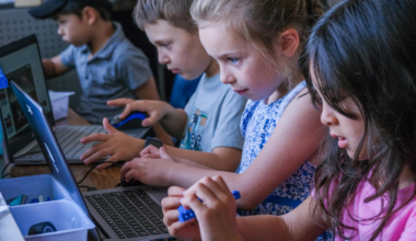 Four children are focused on working with laptops and small electronic components at a table, likely participating in a tech or robotics activity.