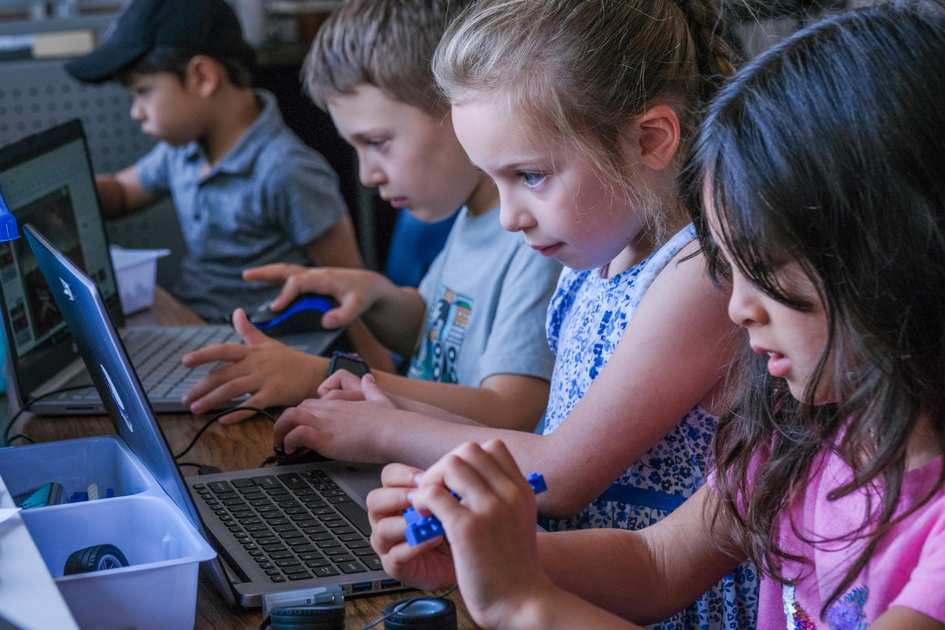 Four children are focused on working with laptops and small electronic components at a table, likely participating in a tech or robotics activity.