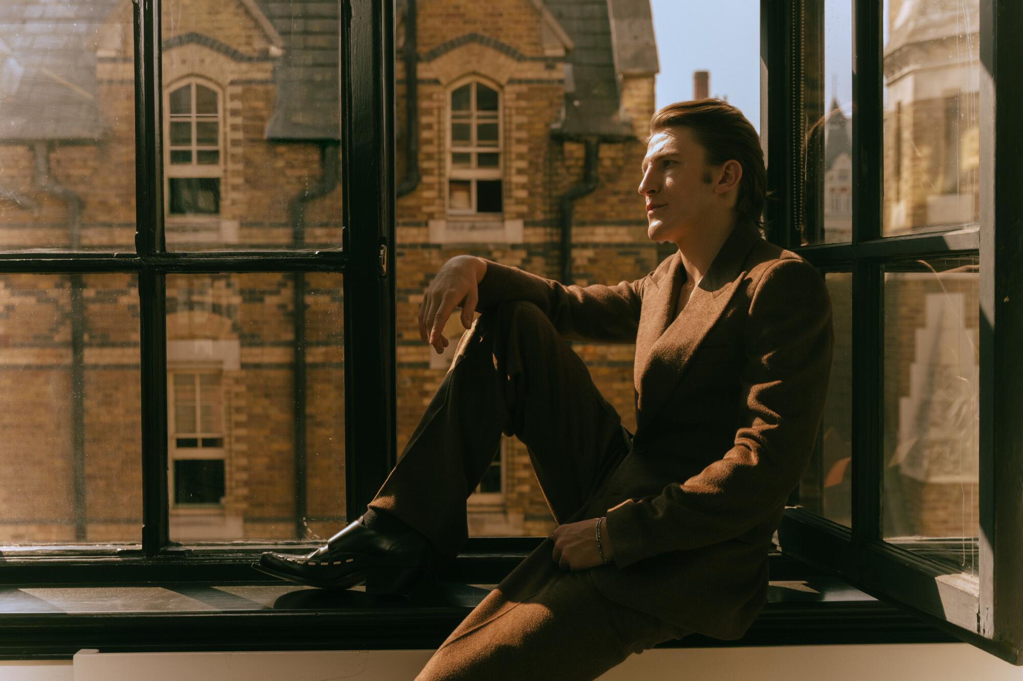 A man in a brown suit leaning against a set of glass windows with his foot propped up on the ledge.