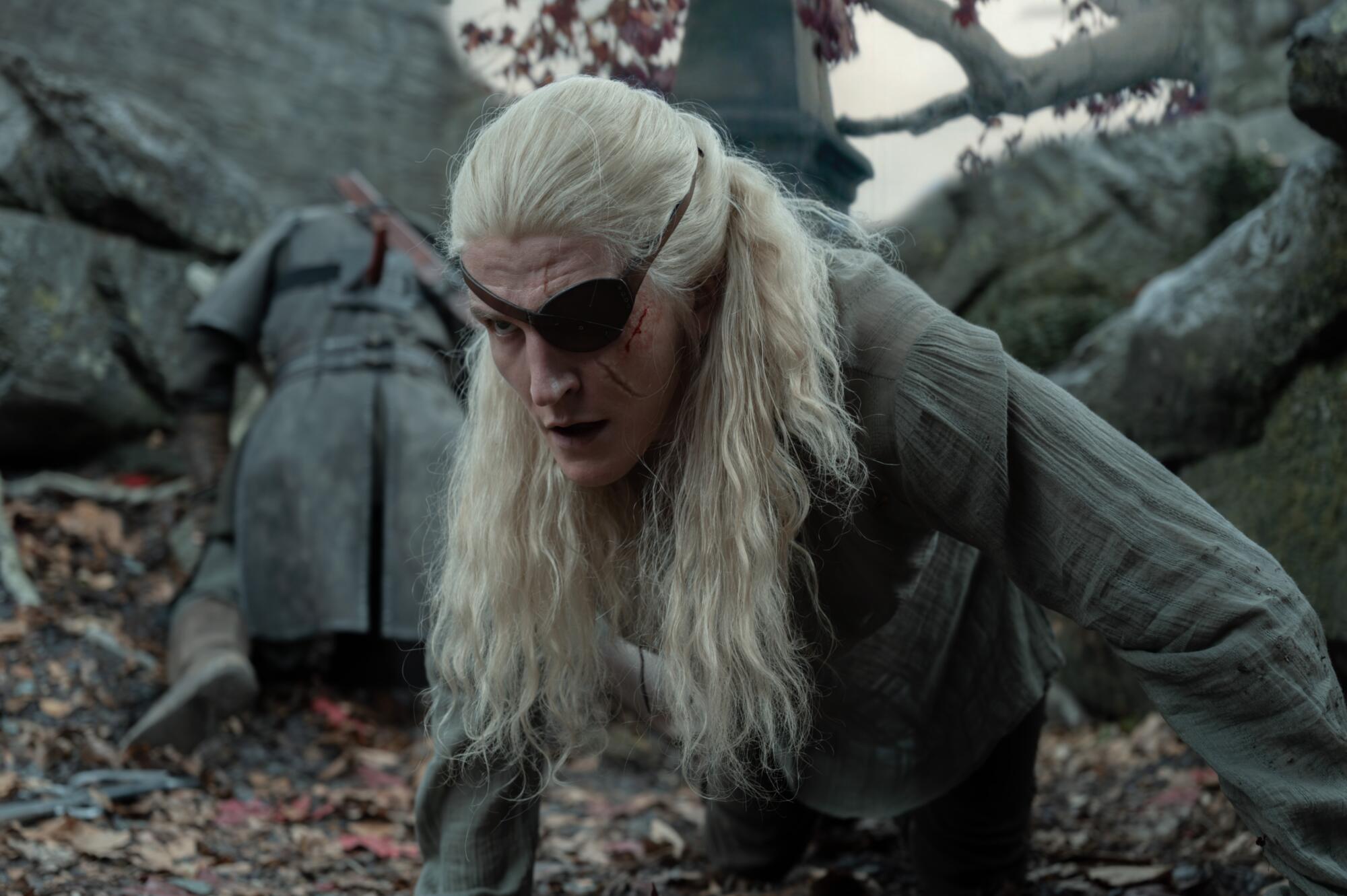 A man with long blond hair and an eye patch with several scratches on his face kneels on the ground.