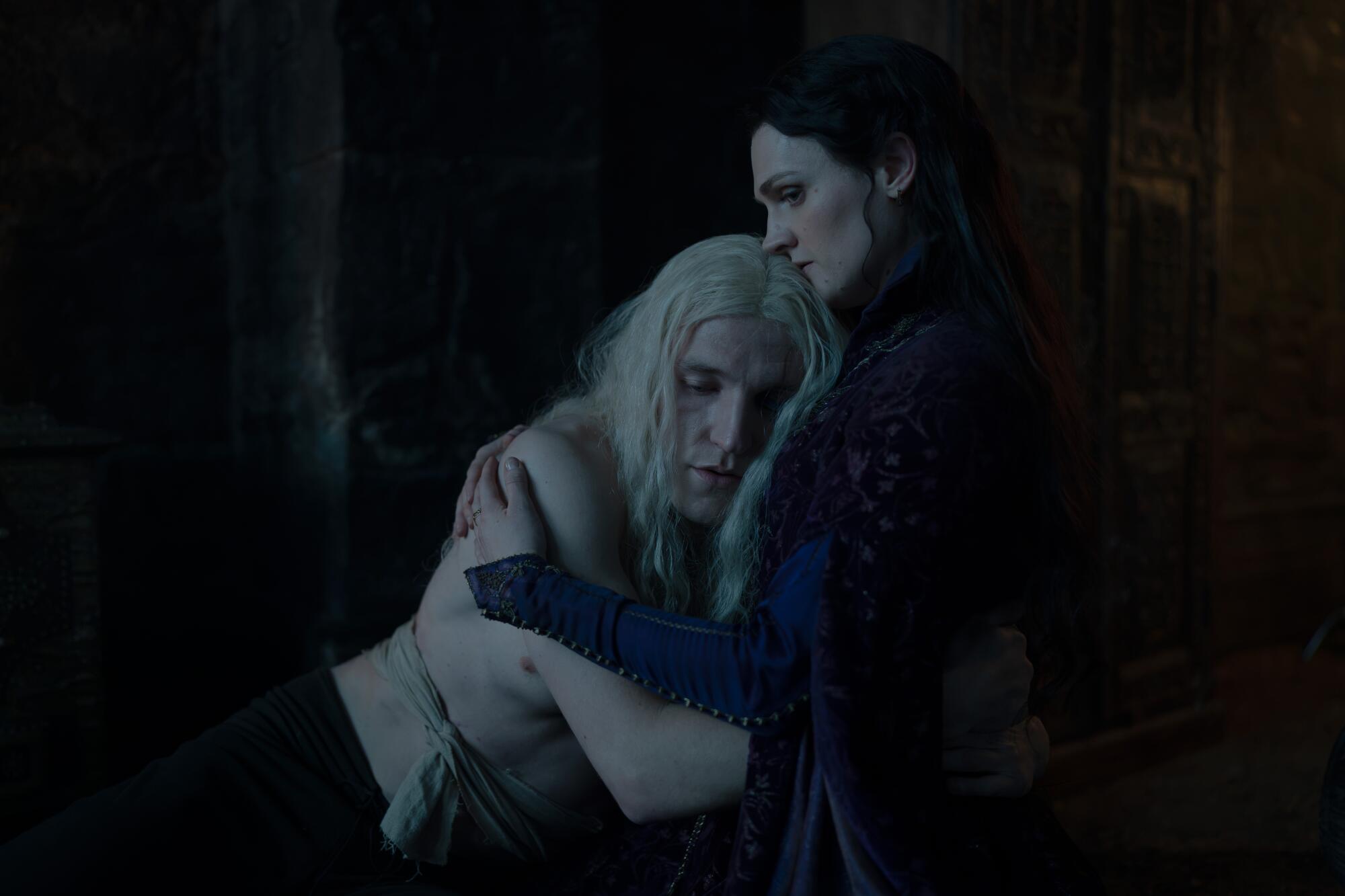 A shirtless man with long blond hair embraces a woman in a dark dress.