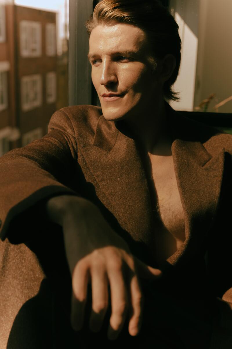 A man in a brown suit with no undershirt looks out a window.