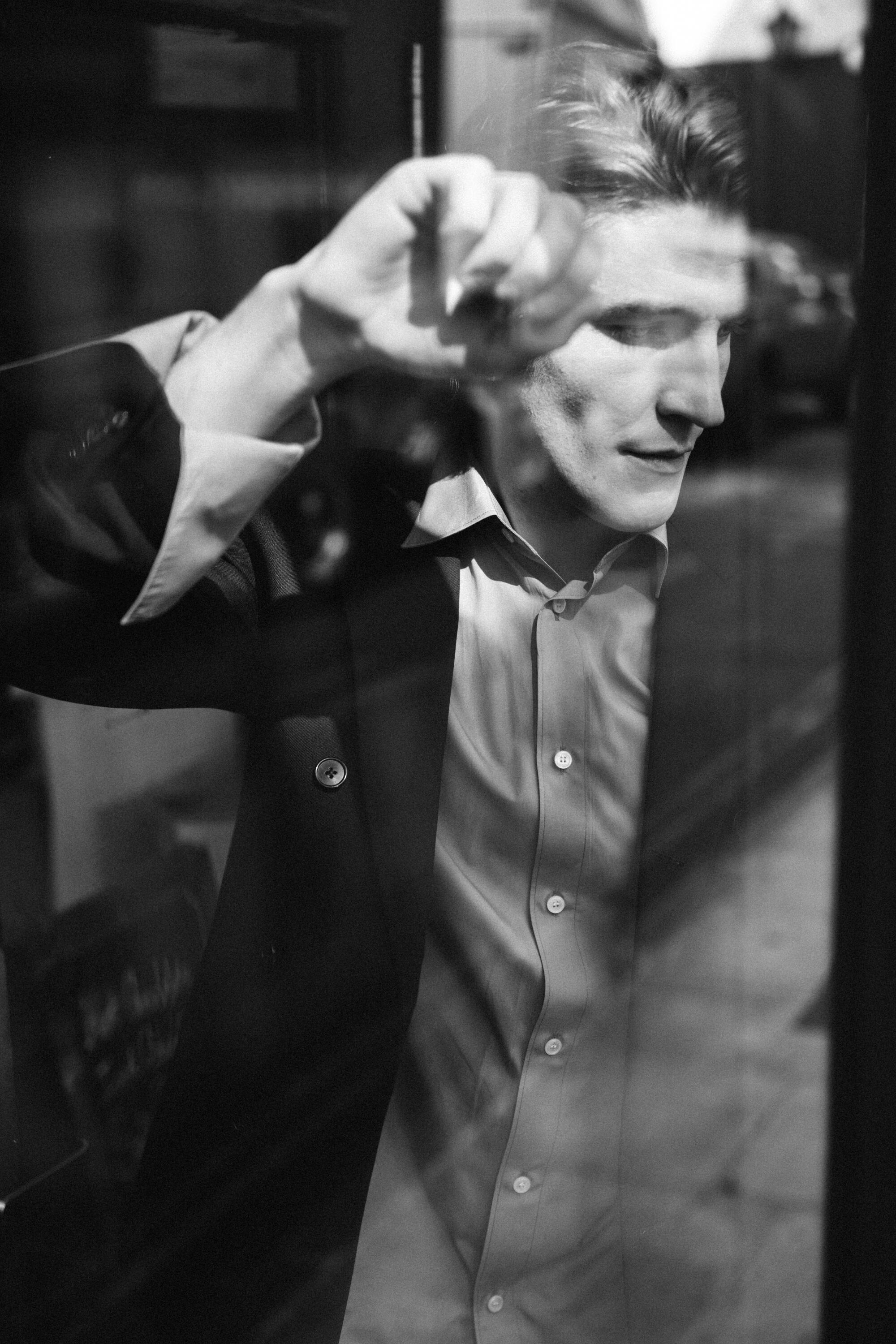 A black and white photo of a man leaning against a window with reflections seen on the glass.