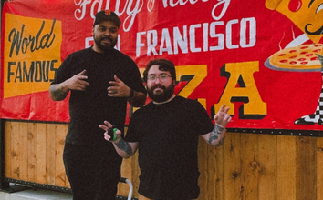Two men pose in front of a red banner reading "Fatty Natty's San Francisco Pizza," with one man standing on crutches smiling and the other with tattoos.