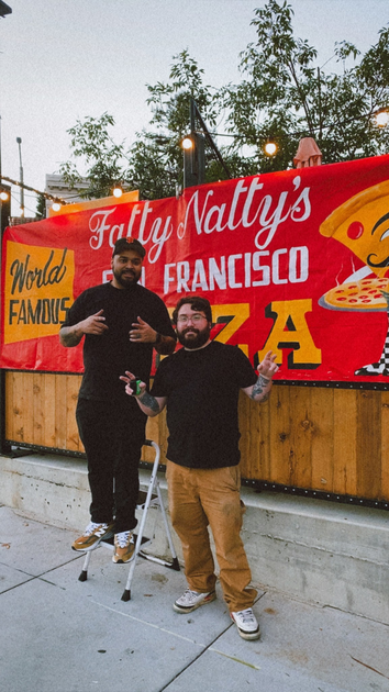 Two men pose in front of a red banner reading "Fatty Natty's San Francisco Pizza," with one man standing on crutches smiling and the other with tattoos.