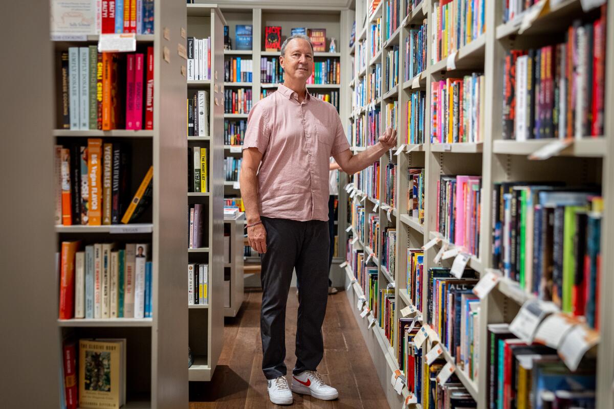 A man with grey hair wearing a pink shirt, grey pants, and white sneakers stands among shelves of books.
