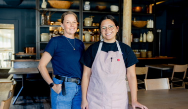 Two women smile inside a warmly lit, rustic restaurant; one wears a navy shirt and jeans, the other a black shirt and a pale pink apron.