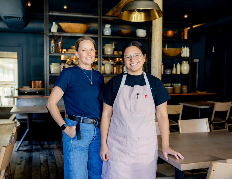 Two women smile inside a warmly lit, rustic restaurant; one wears a navy shirt and jeans, the other a black shirt and a pale pink apron.