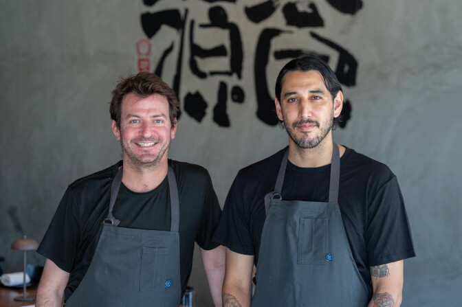 Two chefs in black aprons and t-shirts stand in front of a wall with large Japanese calligraphy. The chef on the left has a light skin tone and brown hair; the chef on the right has a medium-dark skin tone and black hair.