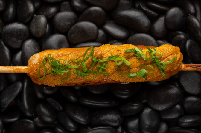 Close-up of a grilled rice skewer topped with glistening uni and scallion, resting on black stones.