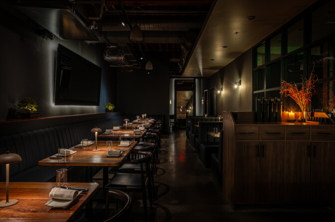 Dark, moody restaurant interior with wood tables, black leather banquette seating, and a hallway lined with warm sconce lighting.