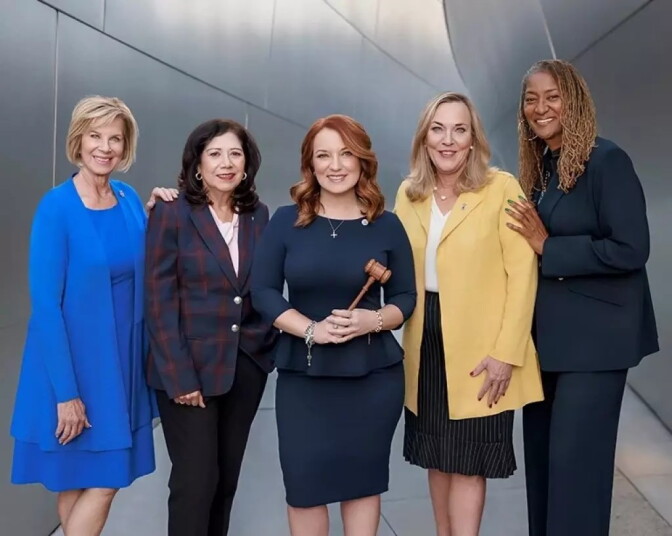 Five women stand and pose for the camera. They are the current L.A. County Supervisors. The woman on the left most side is wearing a blue dress. To her right is a woman wearing a navy blazer with red accents. The woman in the middle holds a gavel and is wearing a dark blue blouse and skirt. On her right, the woman is wearing a long yellow blazer with a white blouse underneath a black skirt. On the right most side, a woman is wearing a dark blue suit.