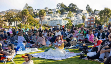 Many people sit on blankets spread over a grassy hill, enjoying a sunny day with buildings and trees in the background.