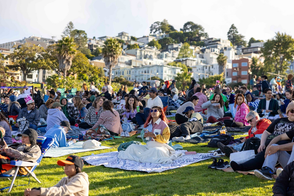 Many people sit on blankets spread over a grassy hill, enjoying a sunny day with buildings and trees in the background.
