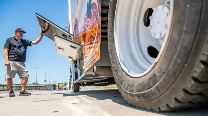 A low angle view of a man, wearing a black polo shirt, shorts, and hat, raising a side door of a food truck. The truck's tire is in the right side of the frame.