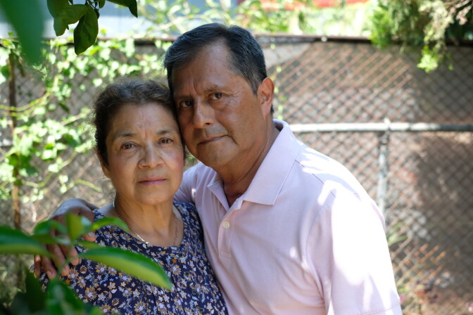 A couple with brown skin tone embrace in a portrait, surrounded by plants. 