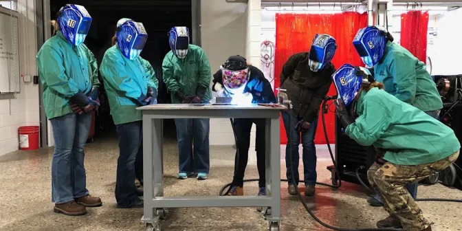 Seveven people wearing protective welding masks and gloves stand around a table while one of them uses a welding torch.