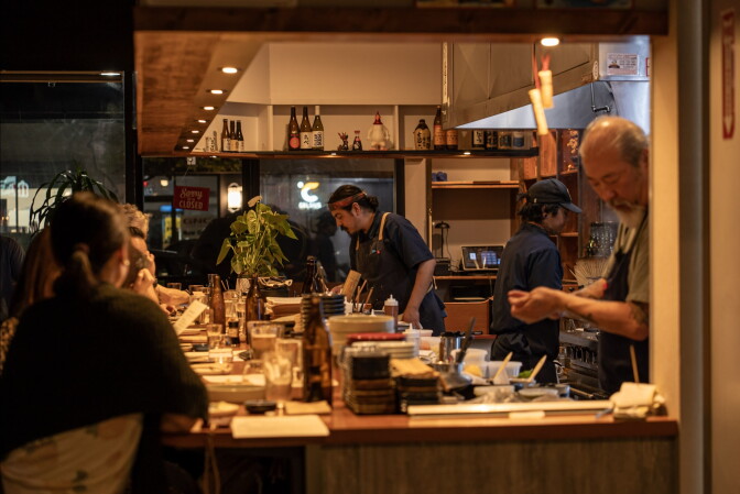 A team of chefs and cooks work behind an open counter as guests look on from the bar.