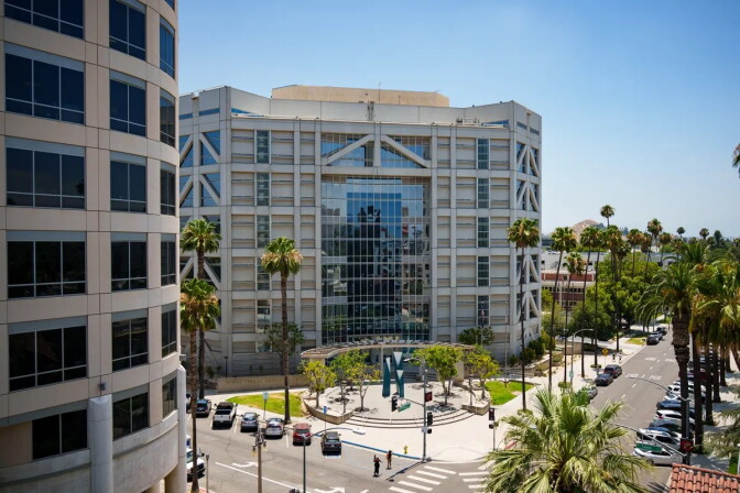 Two multi-story beige buildings. Cars and palm trees are along the sidewalks.