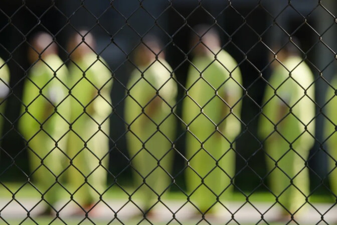 Prisoners in neon yellow jumpsuits are blurred, standing in line. They are pictured through a chain link fence.