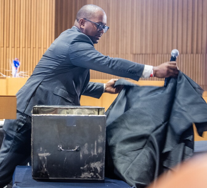 A man wearing a grey suit lifts a black cloth off a worn, steel box with a handle in the middle of its face.