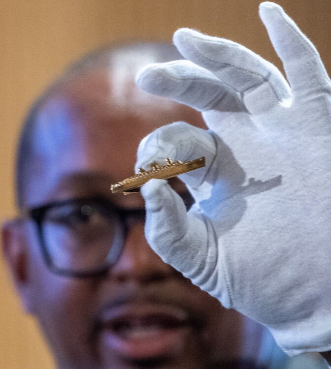 A man holds up a gold tie clip in the shape of a ship. He is wearing a white glove.