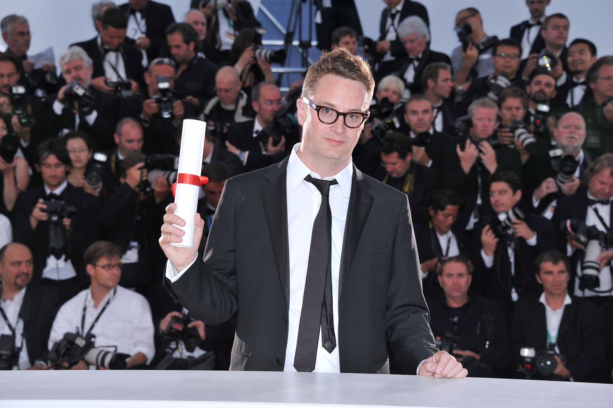 A filmmaker stands in front of press with an award scroll.